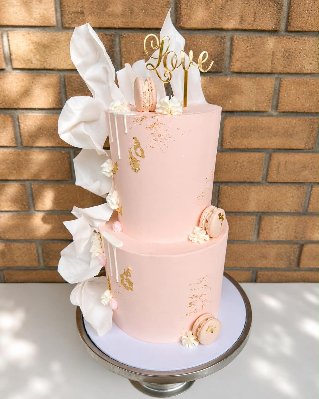 Two tier blush pink wedding cake with white chocolate drip, gold leaf flakes, pink macarons, white wafer paper sails and a gold Love acrylic topper